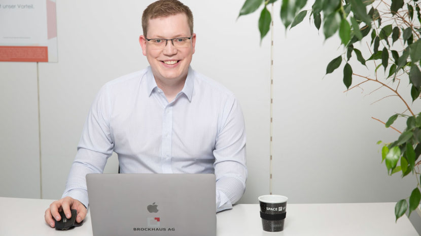 BROCKHAUS-Mitarbeiter David Reher sitzt an einem Schreibtisch, den geöffneten Laptop vor sich